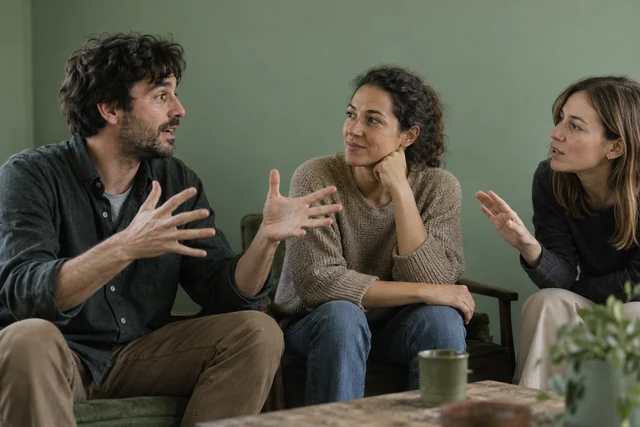 Una persona adulta cuenta una anécdota con manos expresivas a dos oyentes adultos sentados; una persona intenta intervenir con discreción. Escena cotidiana, sin ridiculizar.