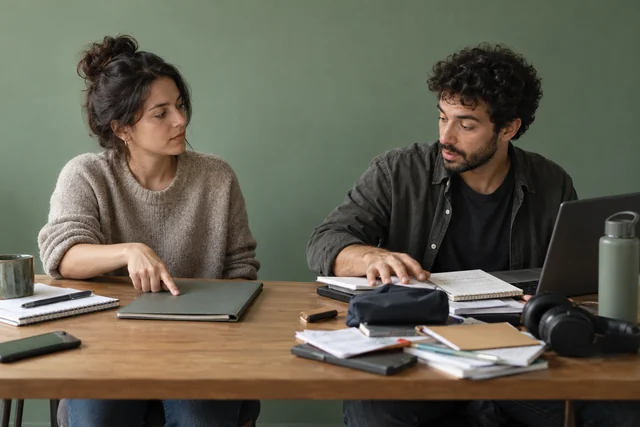 Dos compañeros adultos comparten una mesa; uno señala con calma el borde de su carpeta mientras el otro ha extendido sus objetos por todo el espacio. Gesto contenido de establecer límites.